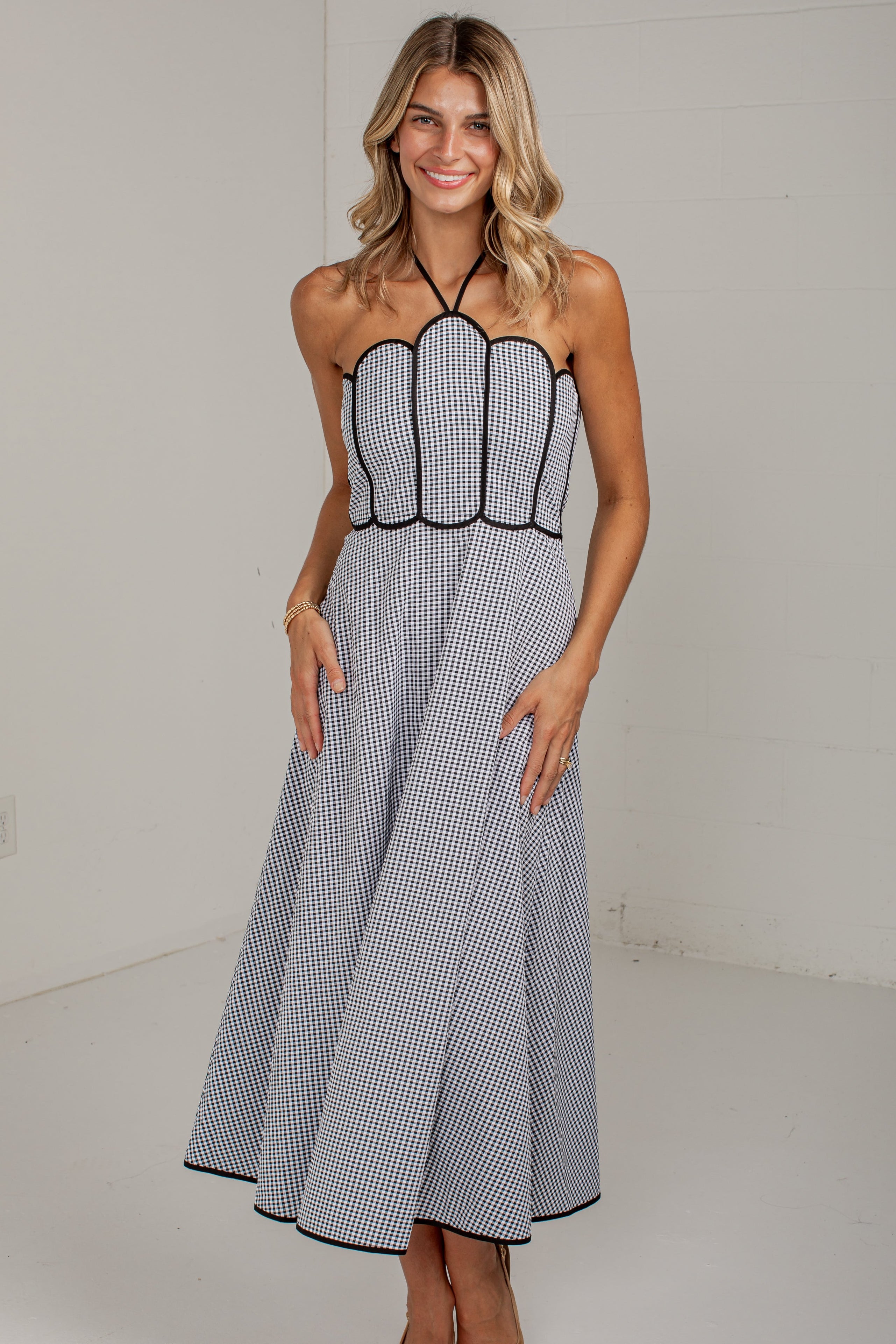 A woman modeling a sleeveless halter-neck midi dress with a black and white gingham pattern and black trim, standing against a plain white studio wall.