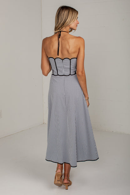 A woman shown from the back wearing a sleeveless midi dress with thin vertical black and white stripes, a scalloped black trim at the waist, and a halter-style back strap, paired with tan heeled sandals in a white studio setting.