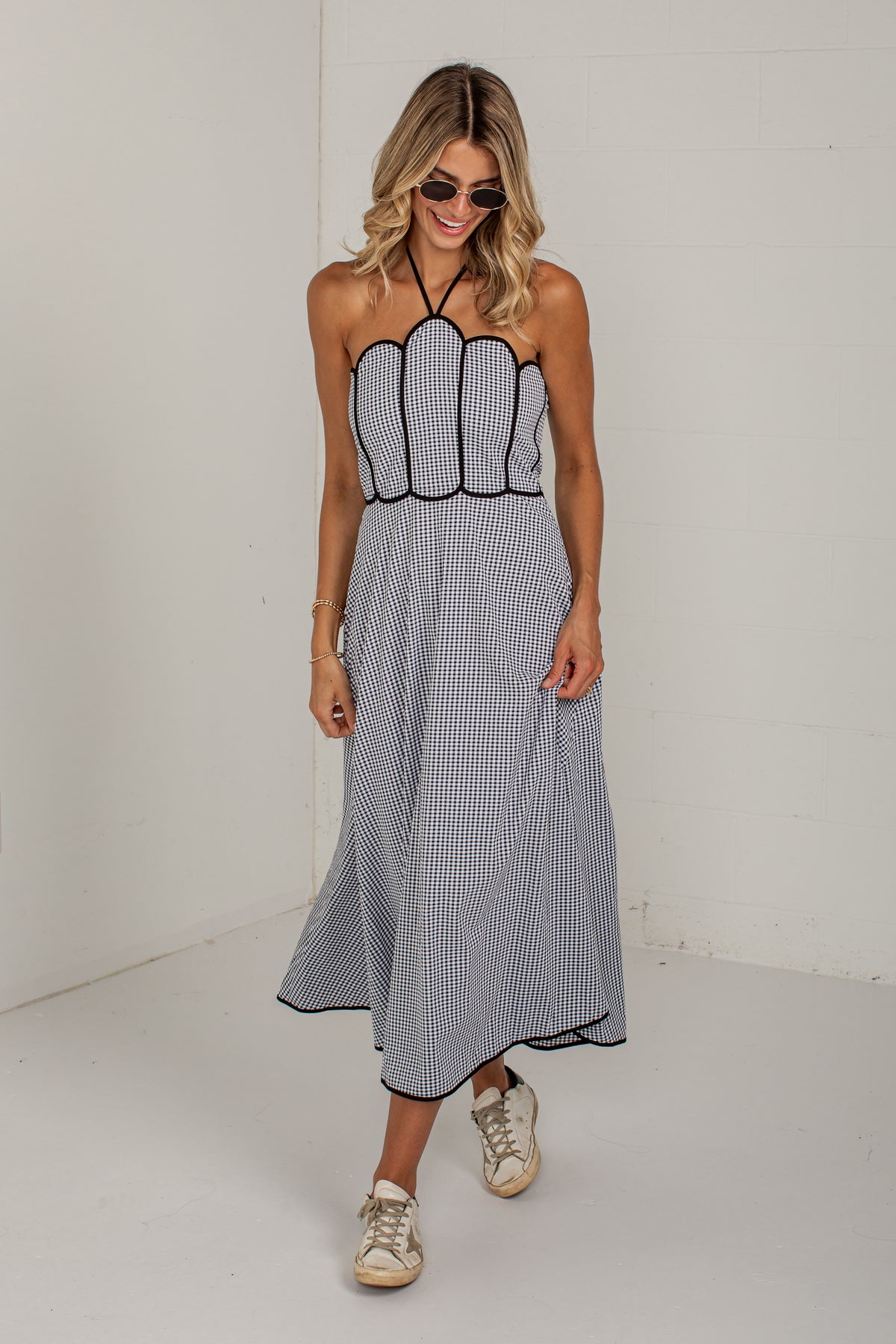 A woman modeling a sleeveless halter-neck maxi dress with a black and white pinstripe pattern, featuring a scalloped bodice and a flowing skirt, paired with beige sneakers in a minimalist white studio setting.