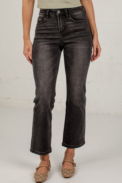 A model wearing a pair of black washed denim jeans with a straight-to-crew leg, paired with a light beige top and brown flat shoes, standing against a white brick wall.