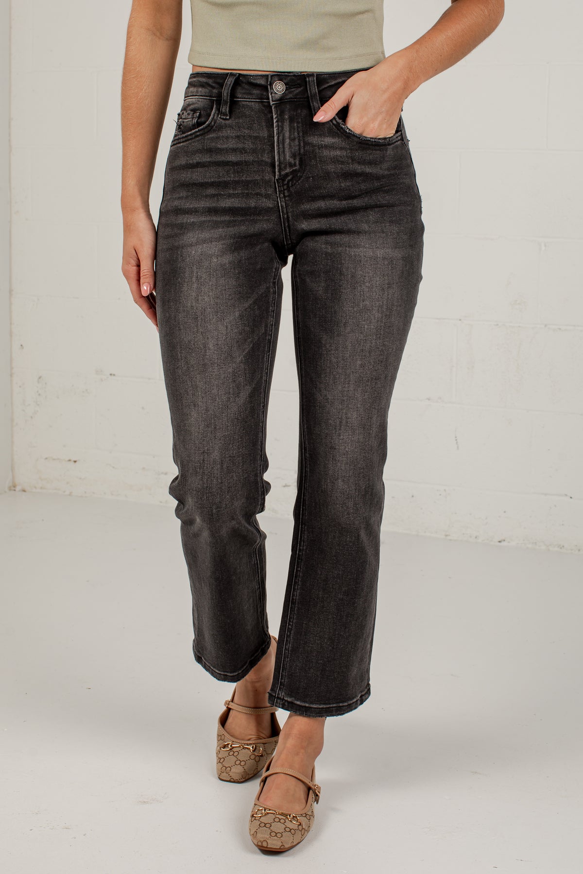A person wearing dark charcoal gray cropped jeans with a faded wash, paired with a beige top and brown flat shoes, standing against a white brick wall.