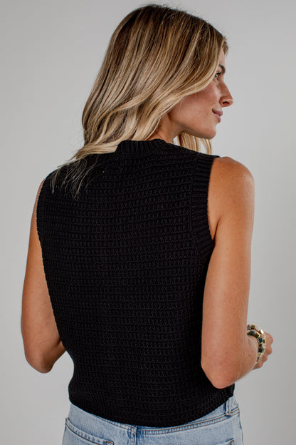 A woman shown from the back wearing a black sleeveless knit top with a textured waffle pattern, paired with light blue jeans, against a plain light gray background.