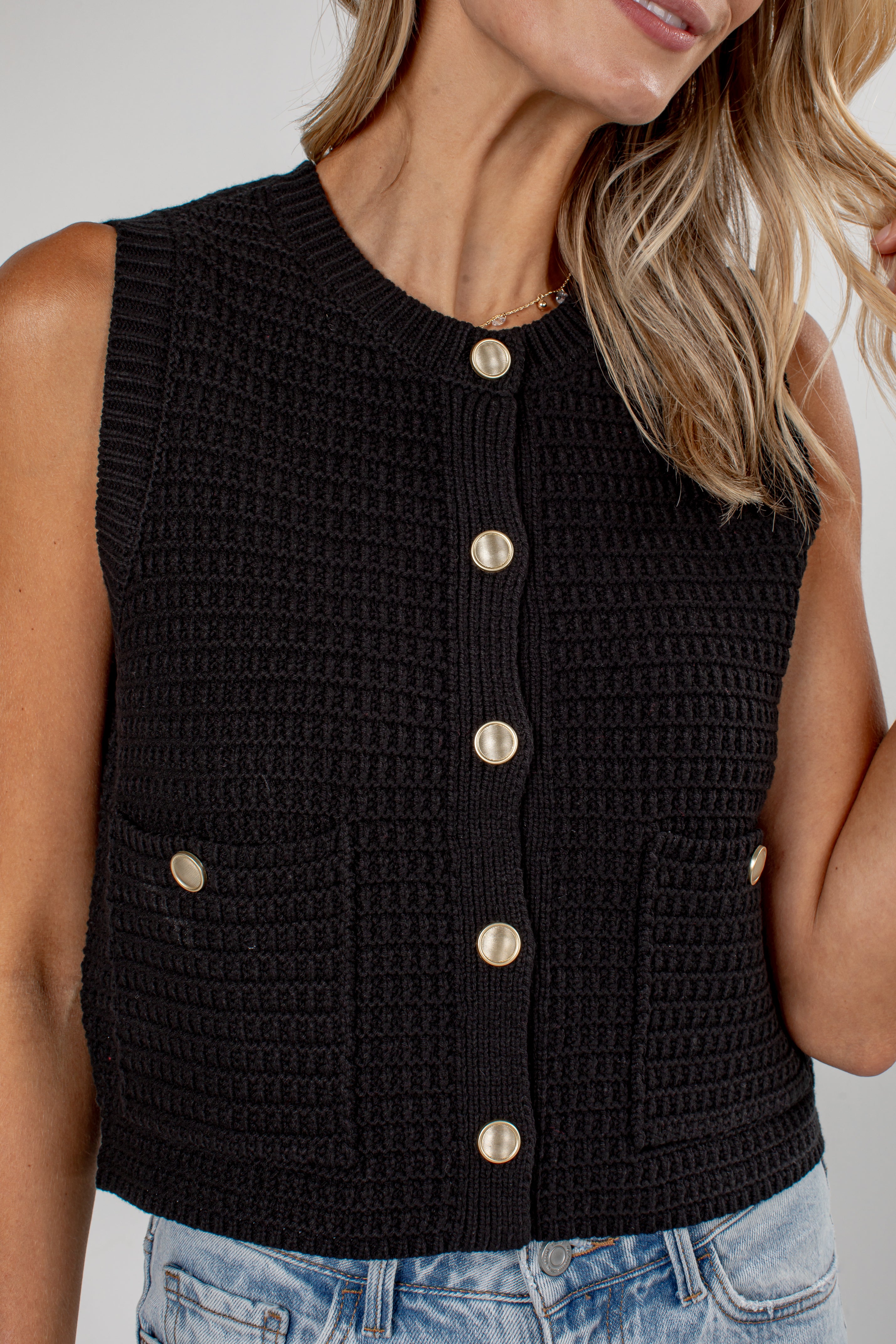 A close-up of a woman wearing a sleeveless black crocheted vest with gold-tone buttons down the front and two matching side pockets, paired with light blue jeans.