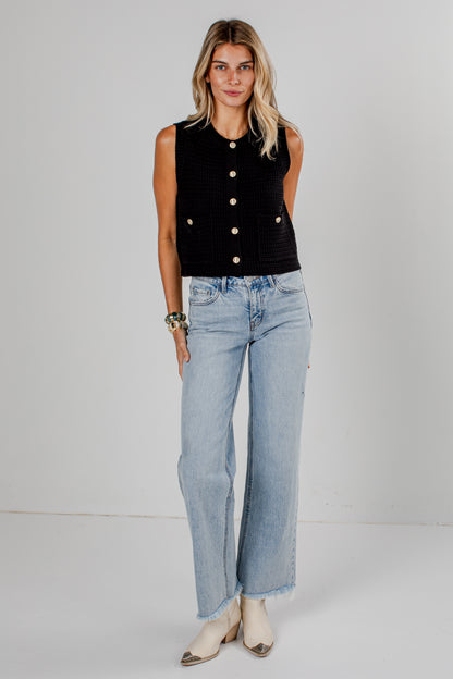 A woman modeling a sleeveless black textured vest with gold buttons and light-wash wide-leg jeans, standing against a plain light gray studio background.