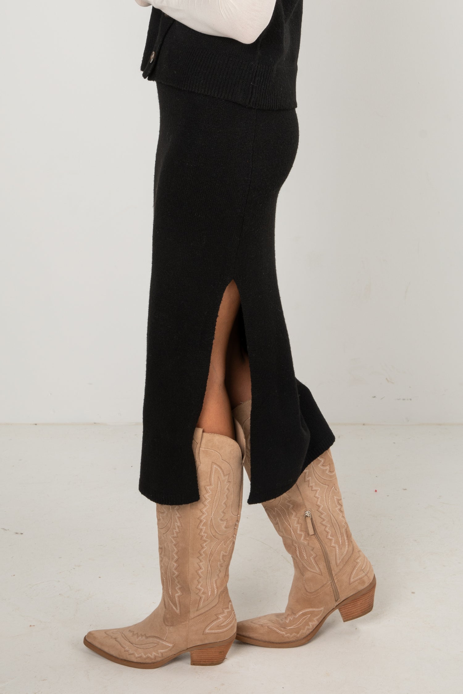 Woman wearing a black knit midi skirt and black buttoned sweater vest layered over a long-sleeve white top, styled with tan western boots, standing in a studio setting.