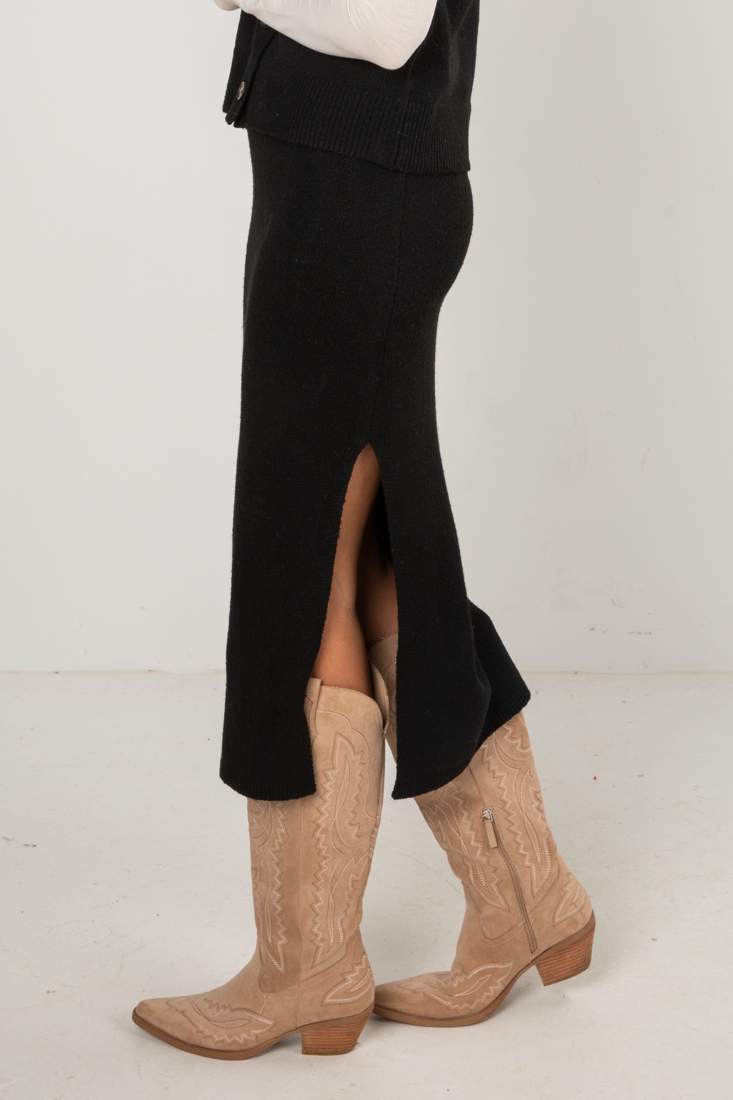 Woman wearing a black knit midi skirt and black buttoned sweater vest layered over a long-sleeve white top, styled with tan western boots, standing in a studio setting.
