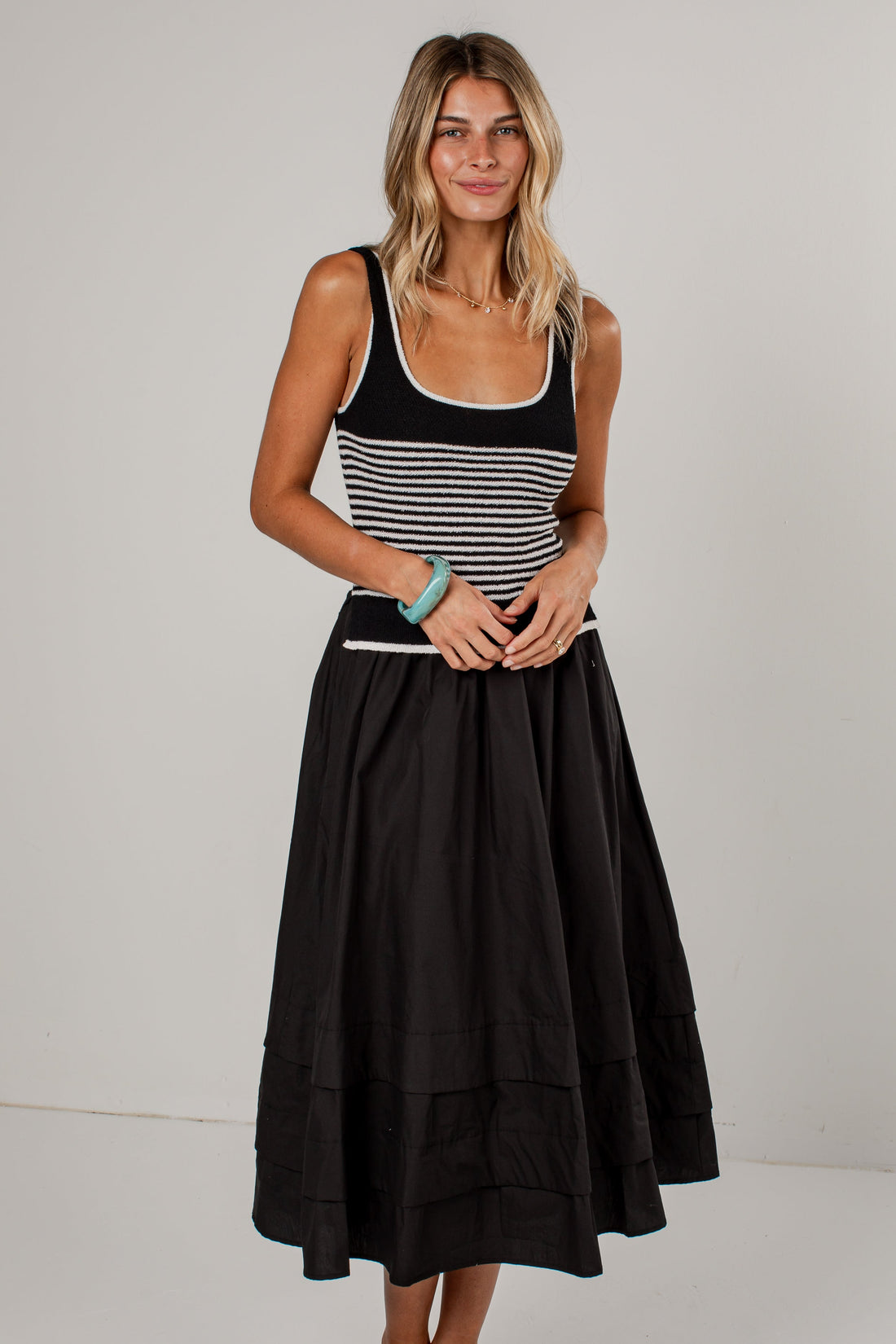 A woman modeling a sleeveless midi dress with a black and white horizontally striped bodice and a full black tiered skirt, paired with tan pointed-toe heels against a plain light gray studio background.
