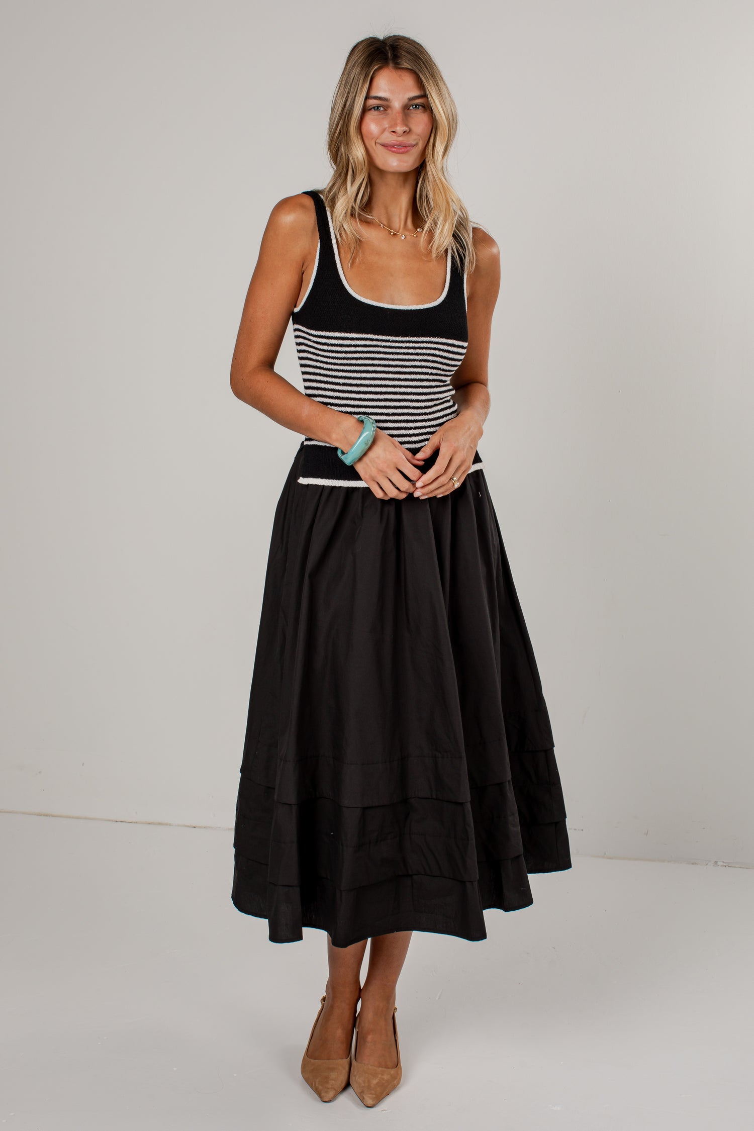 A woman modeling a sleeveless midi dress with a black and white horizontally striped bodice and a full black tiered skirt, paired with tan pointed-toe heels against a plain light gray studio background.