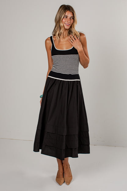 A woman modeling a sleeveless midi dress with a black and white striped bodice and a full black tiered skirt, paired with tan pointed-toe heels against a plain light gray studio background.