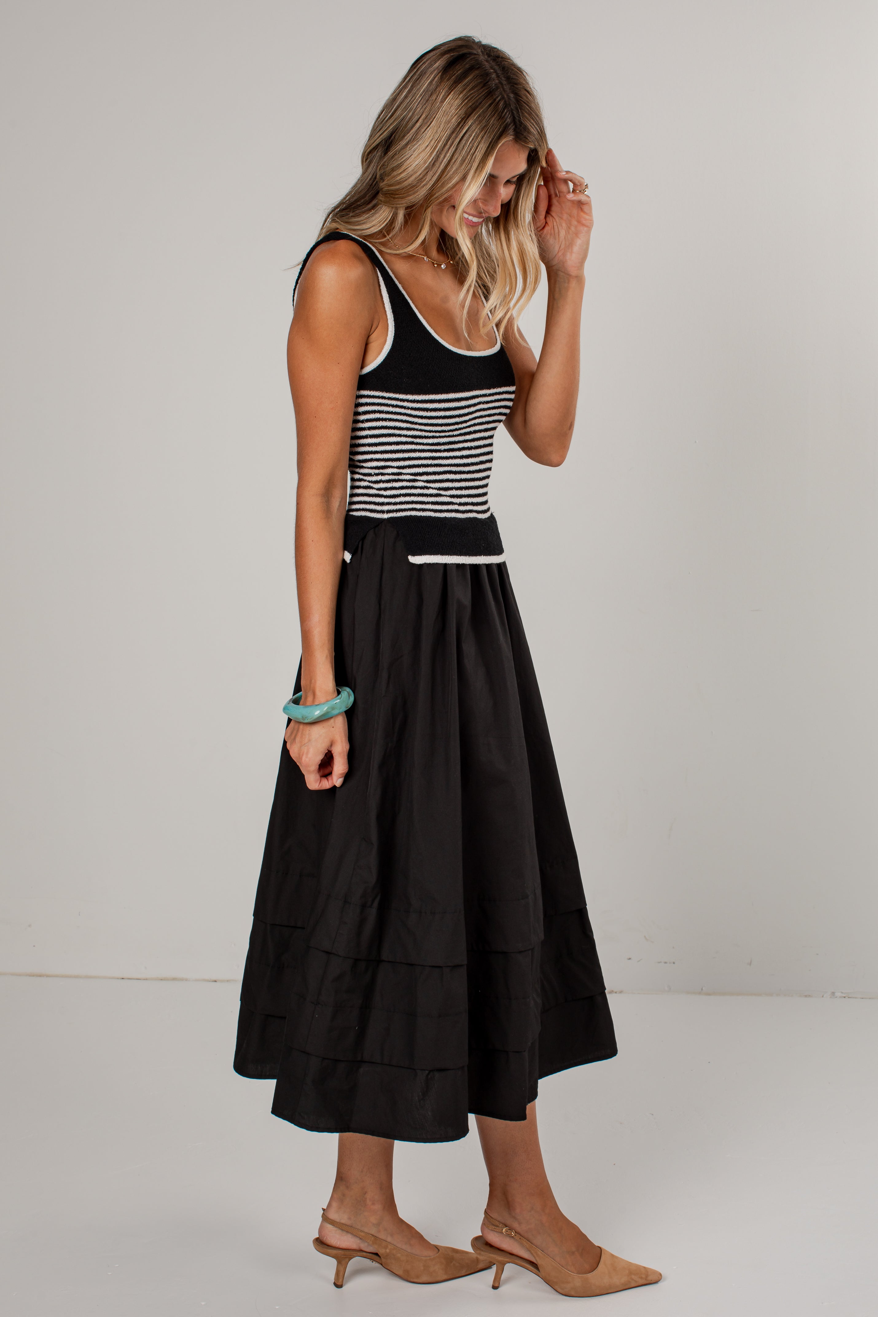 A woman modeling a sleeveless midi dress with a black and white horizontally striped bodice and a full black tiered skirt, paired with tan heeled sandals against a plain light gray studio background.