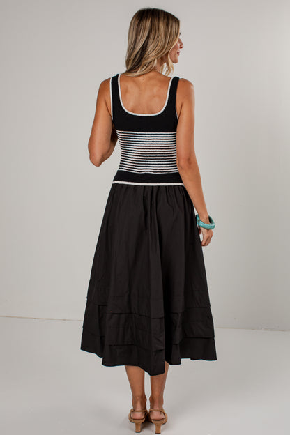 A woman shown from the back wearing a sleeveless black midi dress with a black and white horizontally striped bodice and a full black skirt, photographed against a plain light gray studio background.