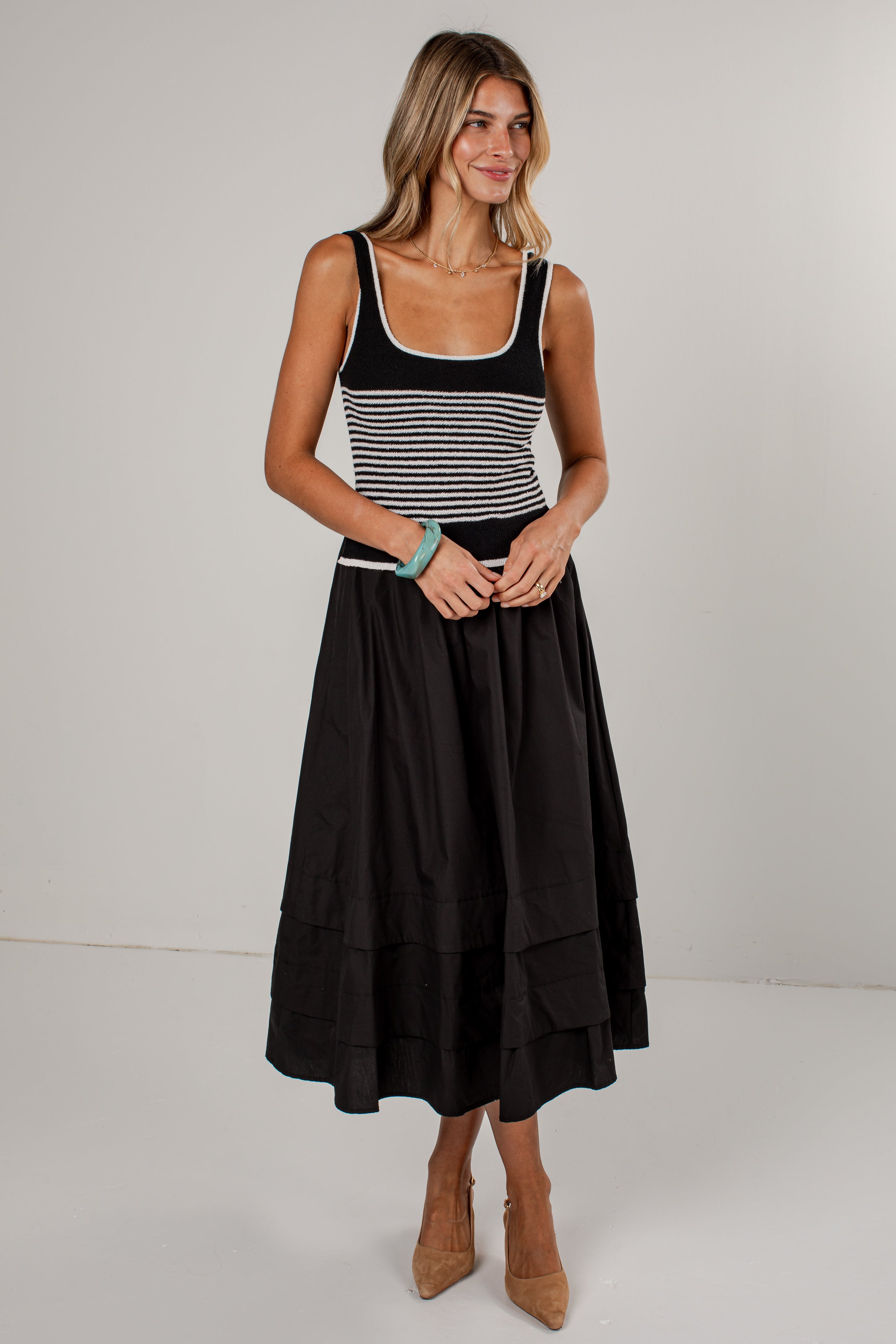 A woman modeling a sleeveless midi dress with a black and white horizontally striped bodice and a solid black tiered skirt, paired with tan pointed-toe heels against a plain light gray studio background.