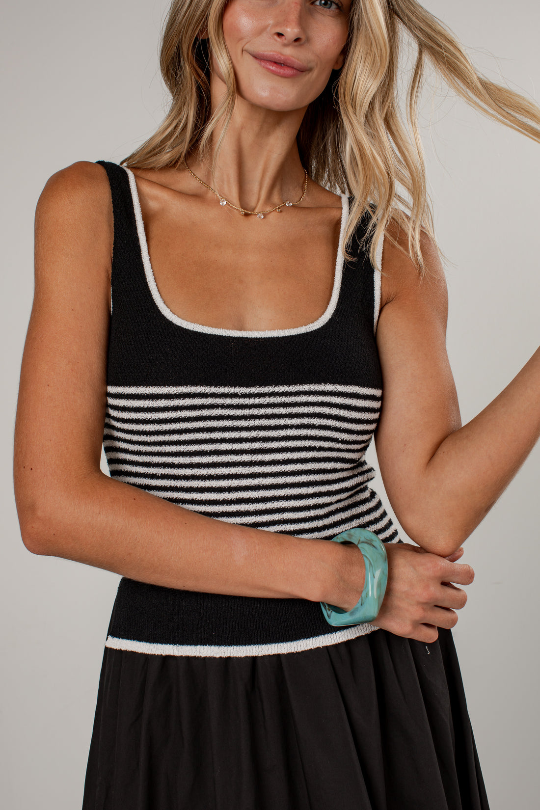 A woman wearing a black knit tank top with white stripes and white trim, paired with a black skirt, against a light gray studio background.