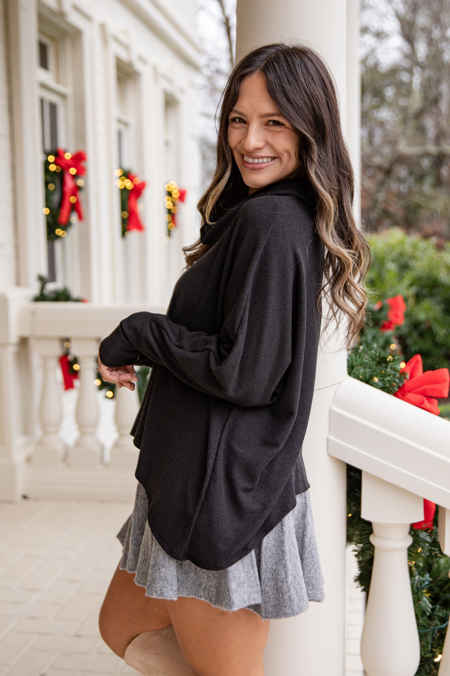 Woman in a black cardigan and gray skirt standing on a porch with Christmas decorations.