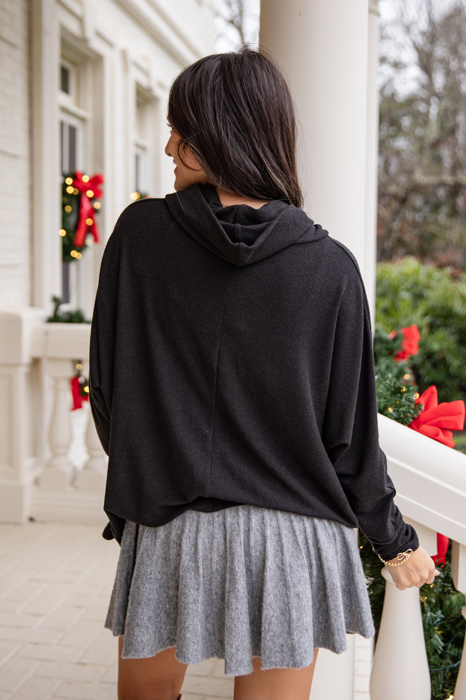 Person wearing a black hoodie and gray skirt on a porch with holiday decorations.