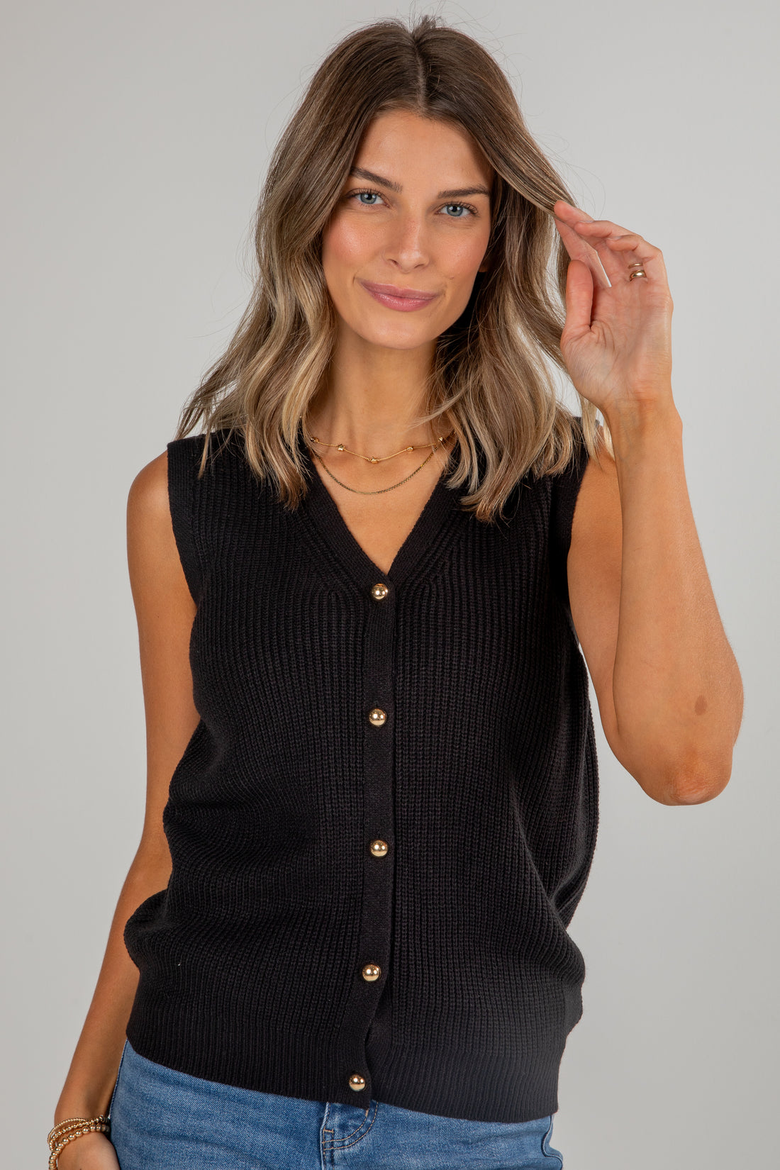 Woman wearing a black sleeveless cardigan with gold buttons on a gray background