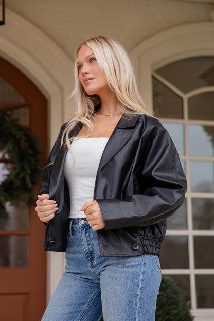 Woman wearing a black jacket over a white top and blue jeans, standing in front of a building entrance.