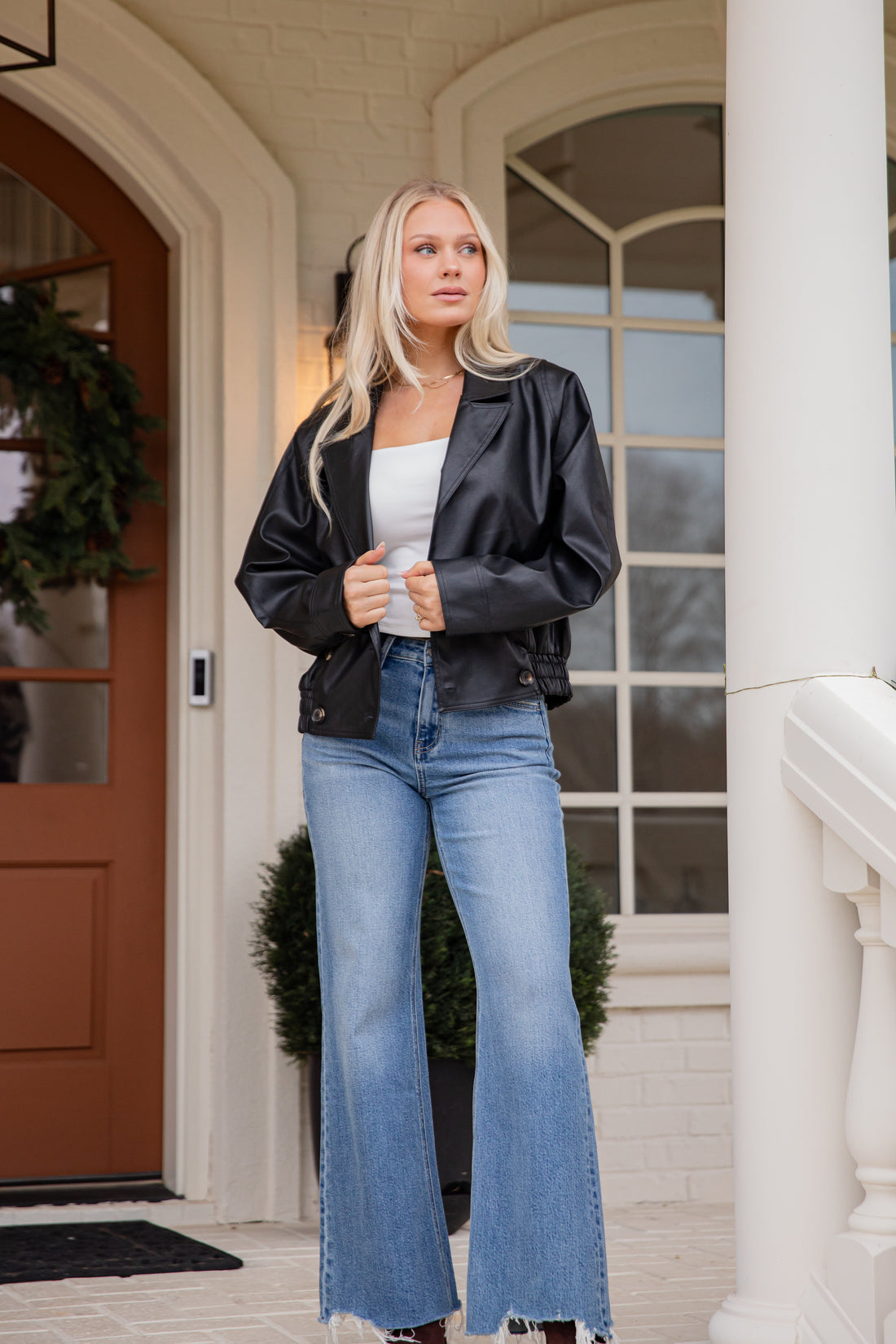 Woman wearing a black jacket, white top, and blue jeans standing outside a house.