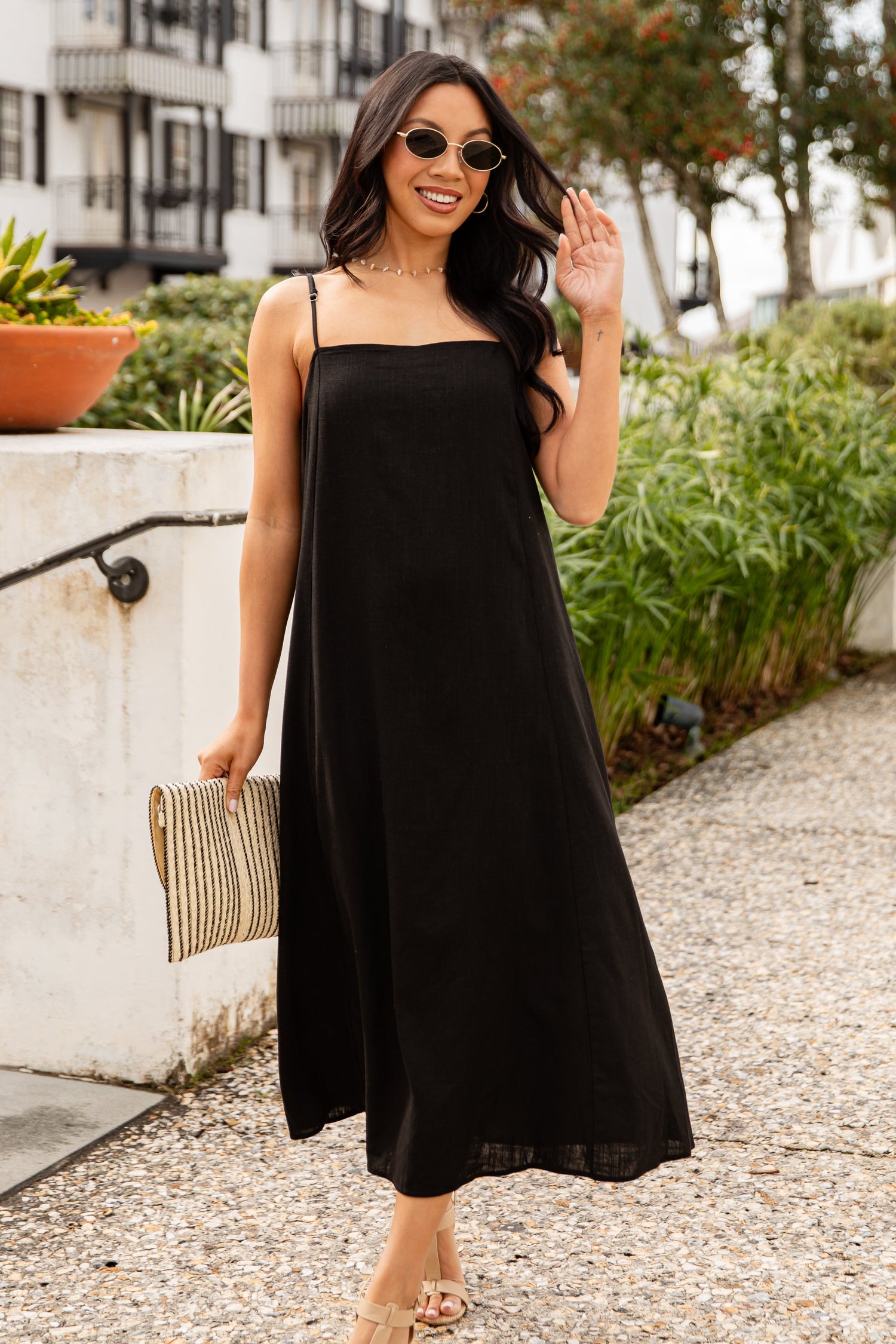 Woman in a black dress standing on a sidewalk with a building and trees in the background