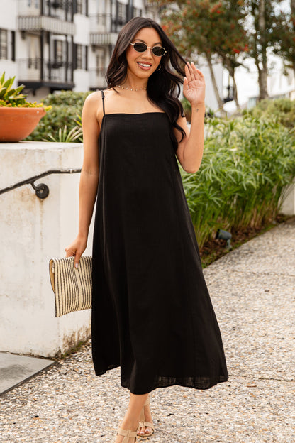 Woman in a black dress standing on a sidewalk with a building and trees in the background