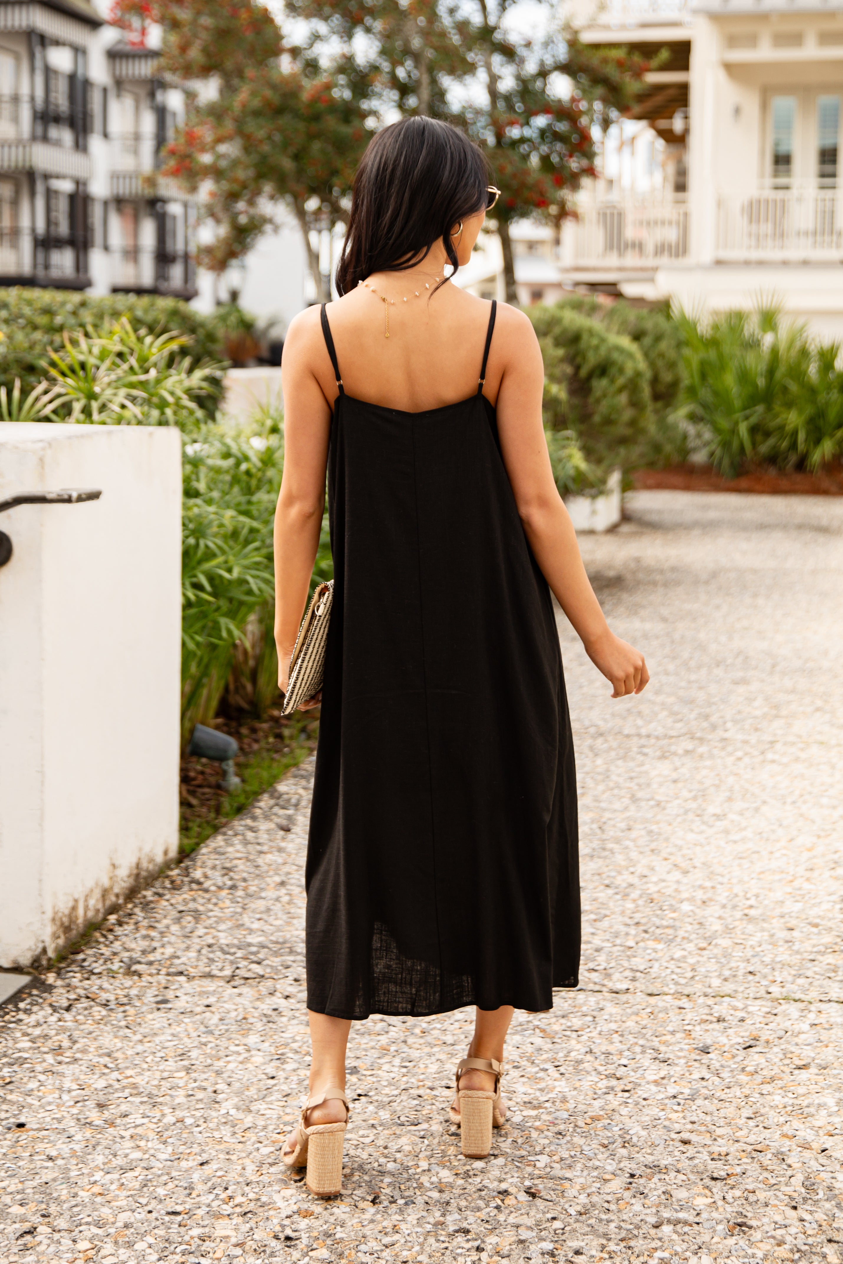 Woman in a black dress walking outdoors with greenery and a building in the background