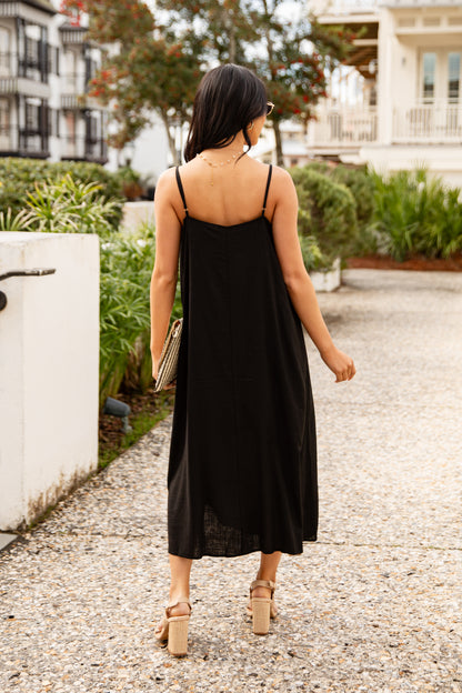 Woman in a black dress walking outdoors with greenery and a building in the background
