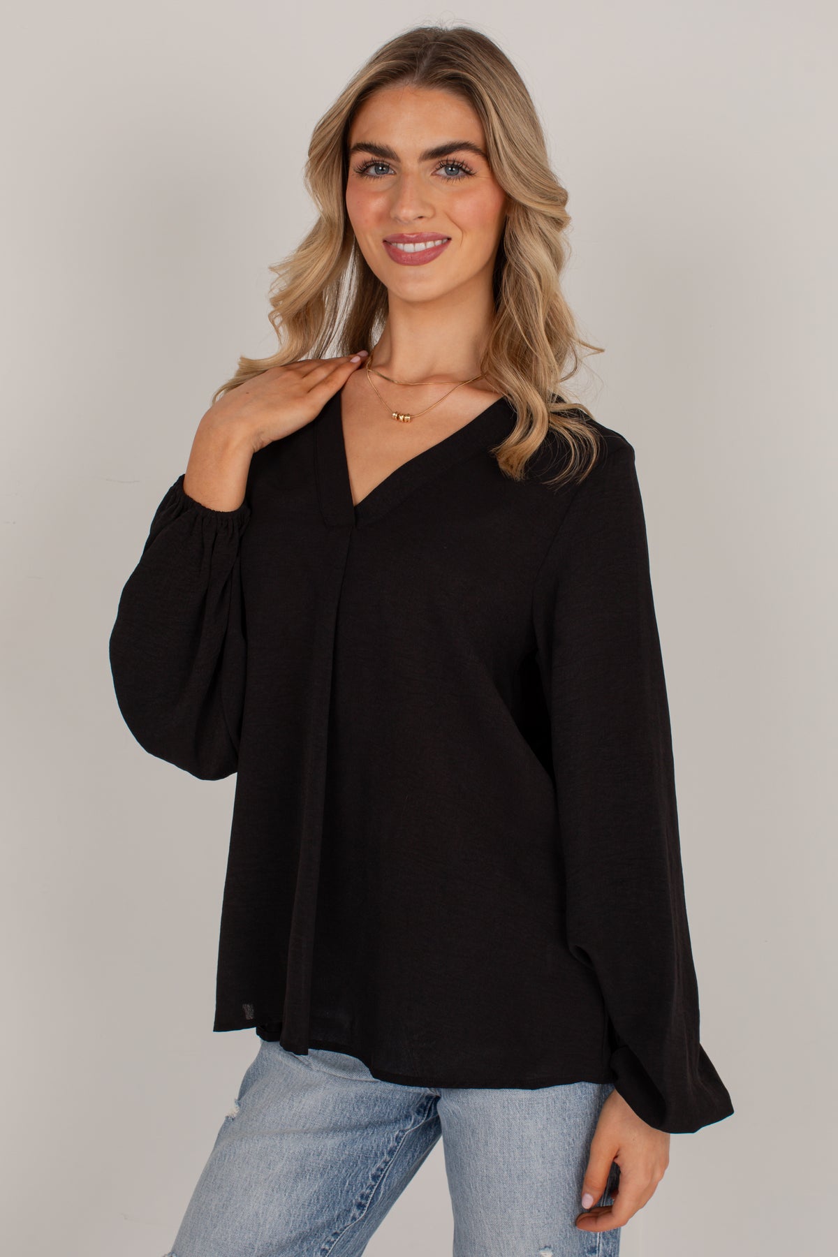 A woman modeling a black long-sleeve blouse with a V-neck and gathered cuffs, paired with light blue jeans, against a plain light gray studio background.