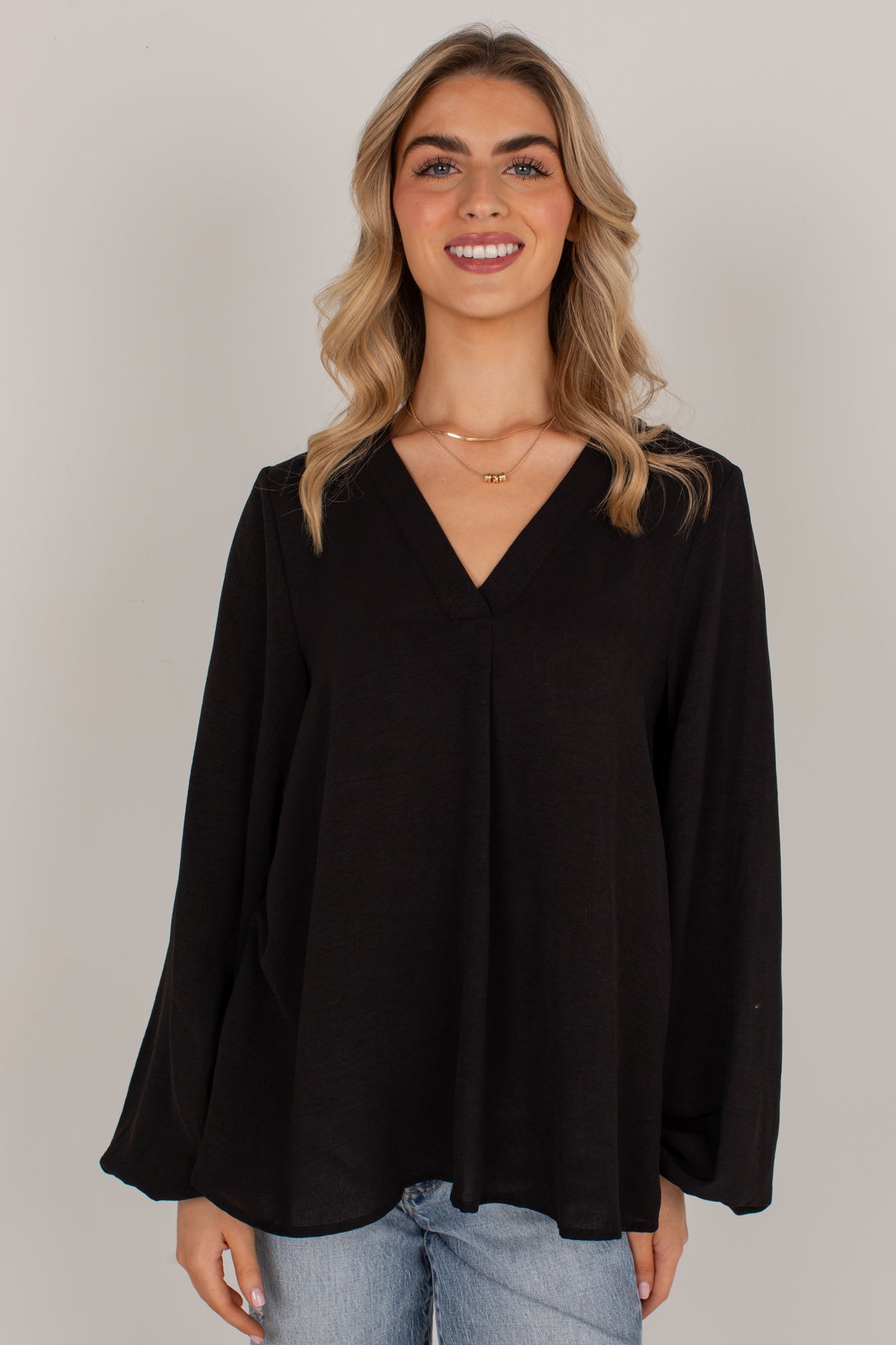 A woman with blonde wavy hair wearing a black long-sleeve V-neck top and light blue jeans, standing against a solid light gray studio background.