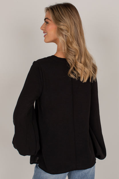 A woman with blonde wavy hair is shown from the back wearing a black long-sleeve top with dramatic bell sleeves and light blue jeans, set against a plain light gray studio background.