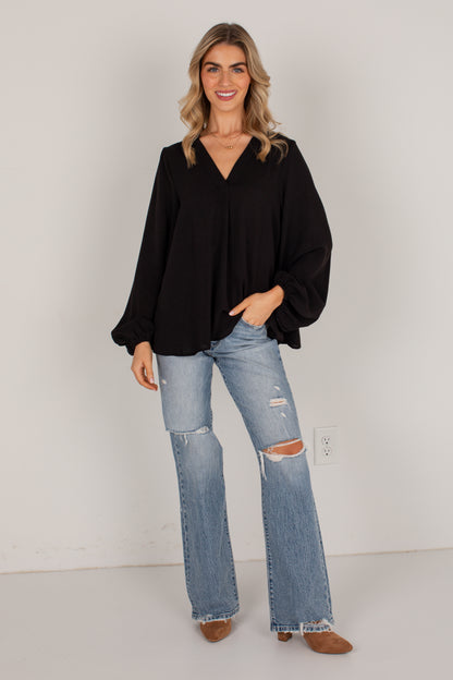 A woman modeling a black long-sleeve blouse with a V-neck and balloon sleeves, paired with light-wash distressed flare jeans and brown shoes, standing against a plain light gray wall.