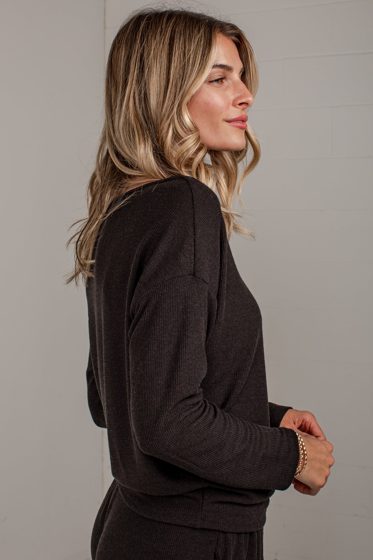 A woman with blonde-highlighted hair is shown in profile wearing a dark brown long-sleeve knit top and matching bottoms against a light gray wall.
