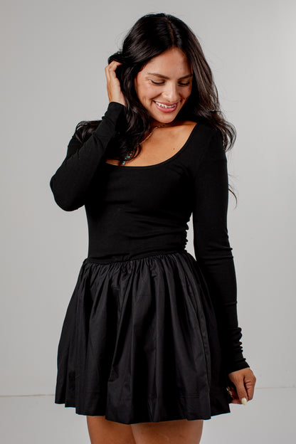 A woman wearing a black long-sleeve scoop-neck top and a black pleated mini skirt, posing against a light gray studio background.