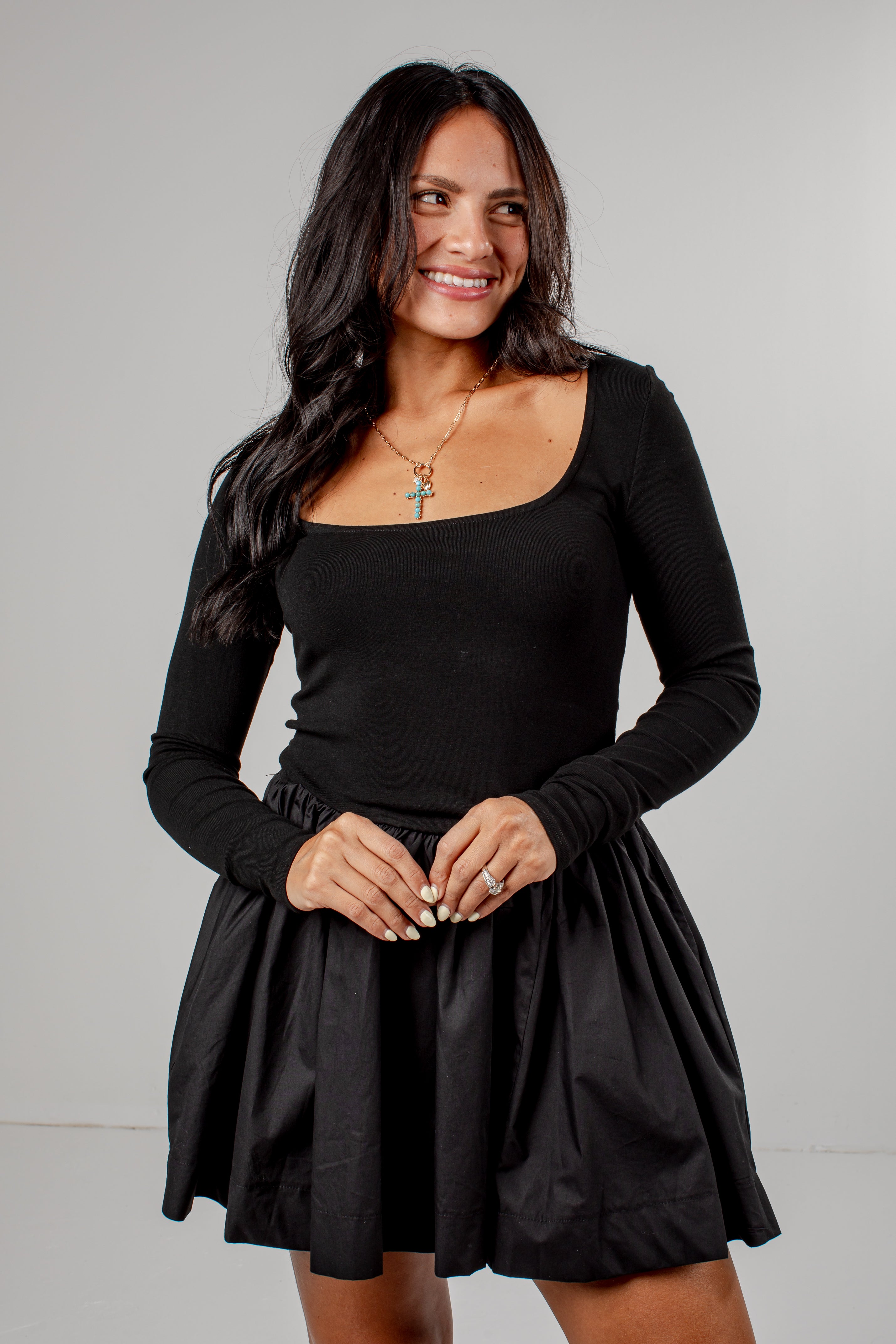A woman modeling a black long-sleeve scoop-neck top with a matching black pleated mini skirt, photographed against a neutral light gray studio background.