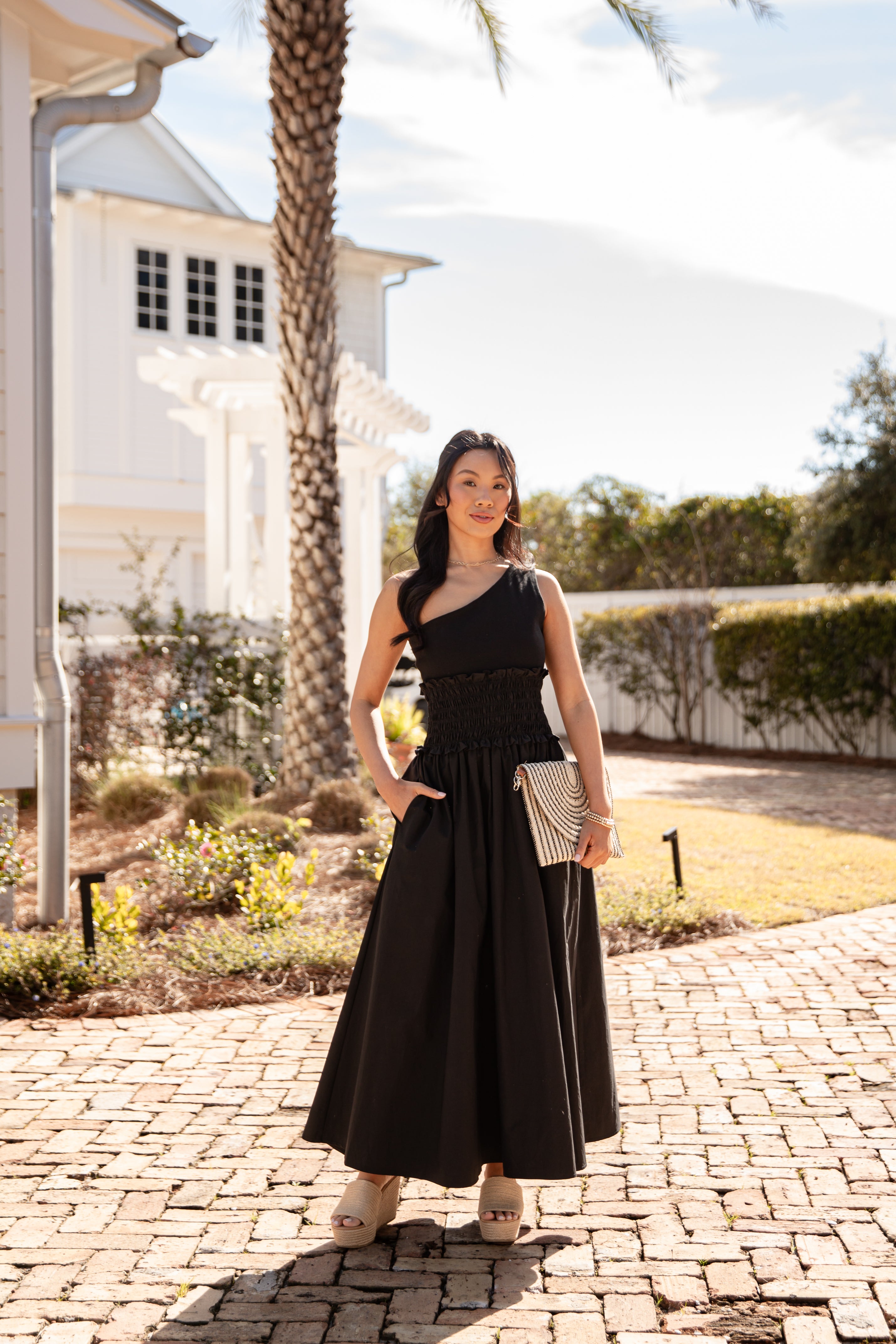Woman in a black dress standing on a paved driveway with a house and palm tree in the background.