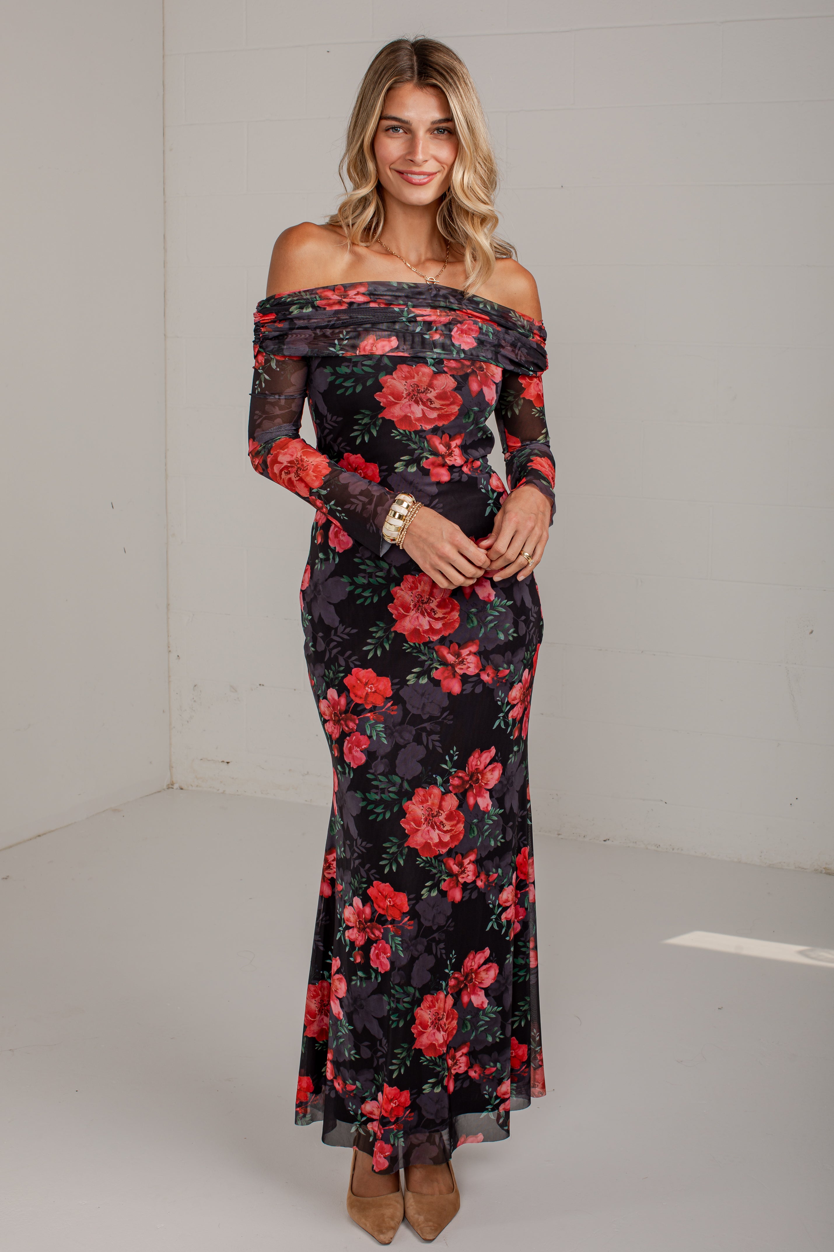 A woman modeling an off-the-shoulder long-sleeve maxi dress with a red and pink floral print on a black background, standing in a white studio corner.