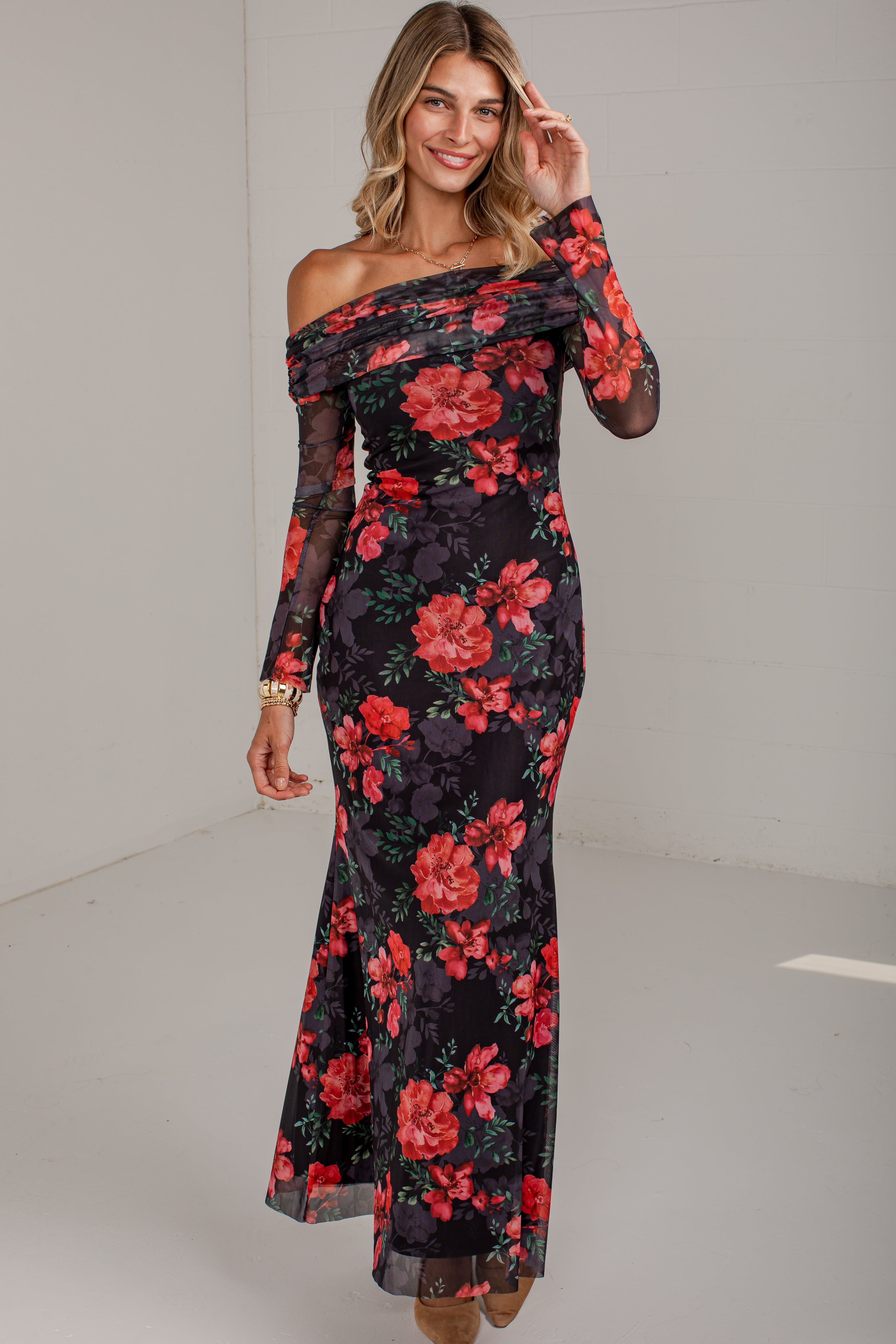 A woman modeling a long off-the-shoulder maxi dress with a black base and a vibrant red and pink floral print, standing in a studio setting.