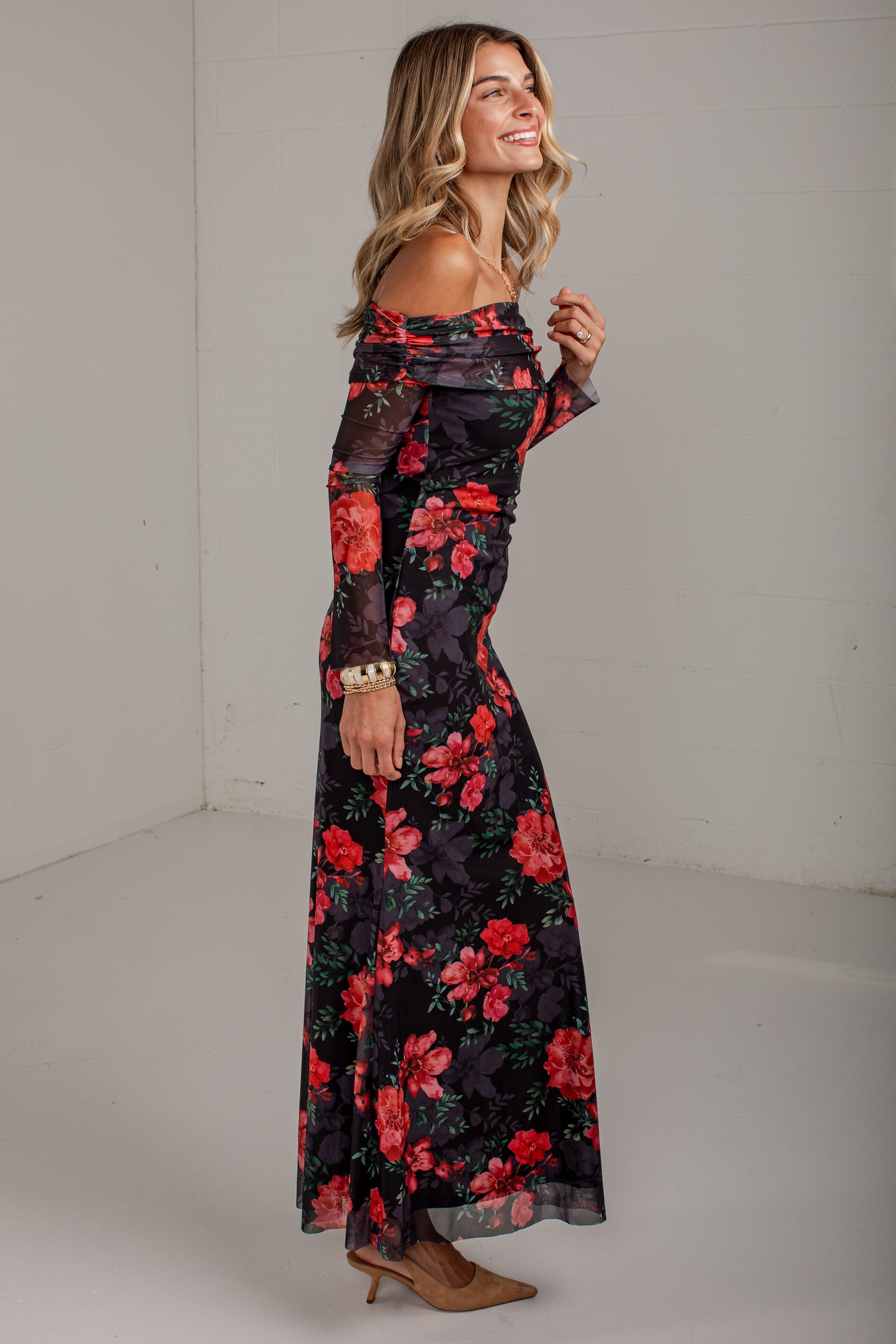 A woman modeling a long off-the-shoulder maxi dress with a black base and a vibrant red and pink floral print, standing in a plain white studio corner.