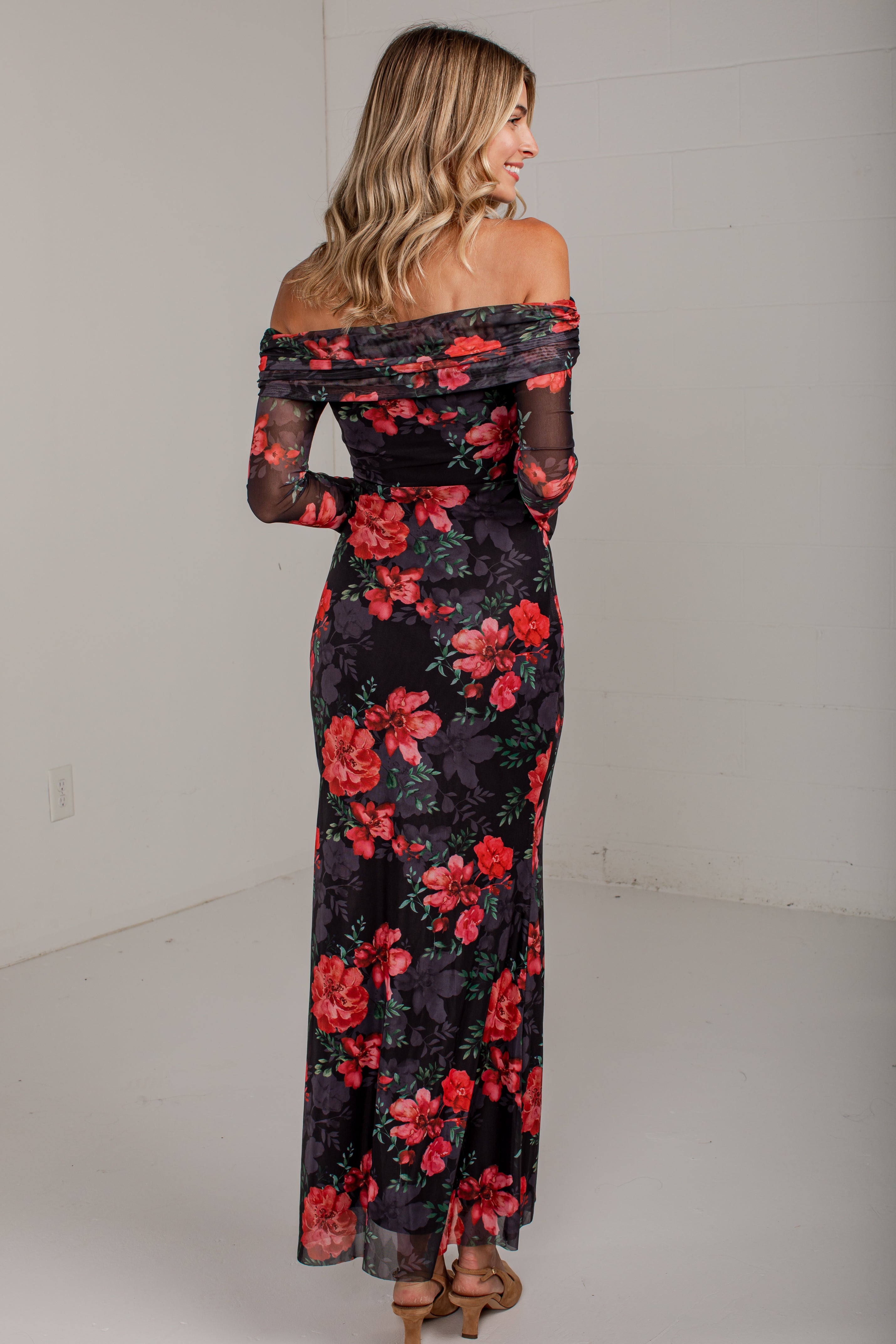 A woman wearing an off-the-shoulder black maxi dress with a vibrant red and pink floral print, photographed from the back in a minimalist white studio setting.