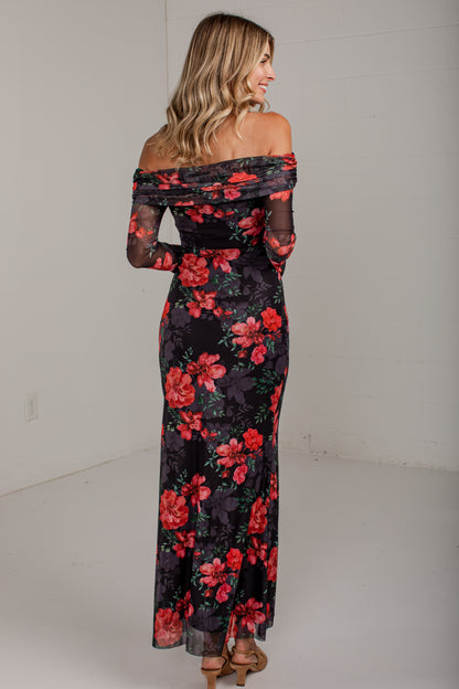 A woman wearing an off-the-shoulder black maxi dress with a vibrant red and pink floral print, photographed from the back in a minimalist white studio setting.