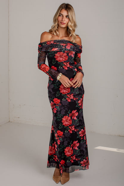 A woman modeling an off-the-shoulder black maxi dress with a vibrant red and pink floral print, standing against a plain light gray wall.