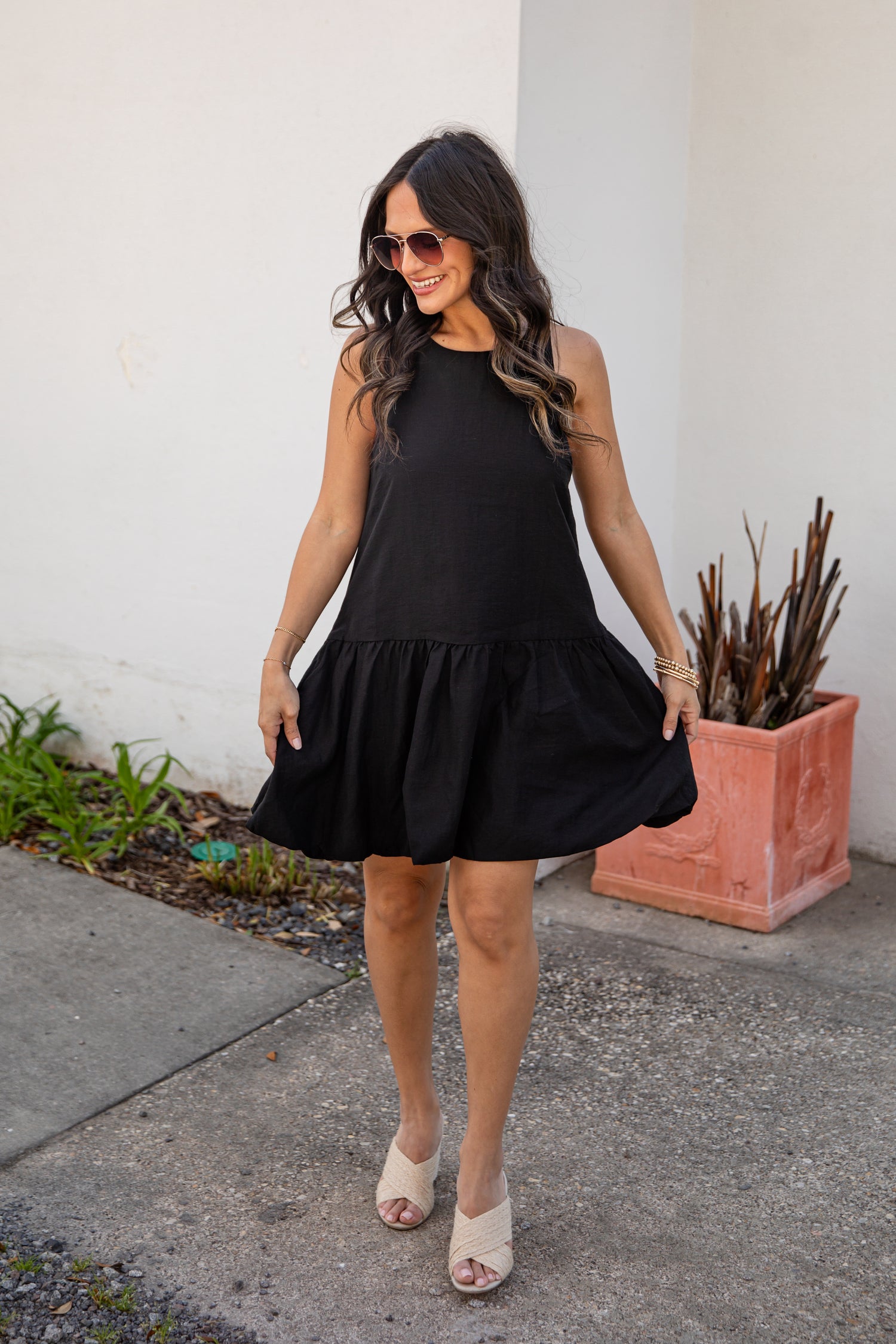 Woman wearing a black dress standing outdoors next to a white wall and potted plant.