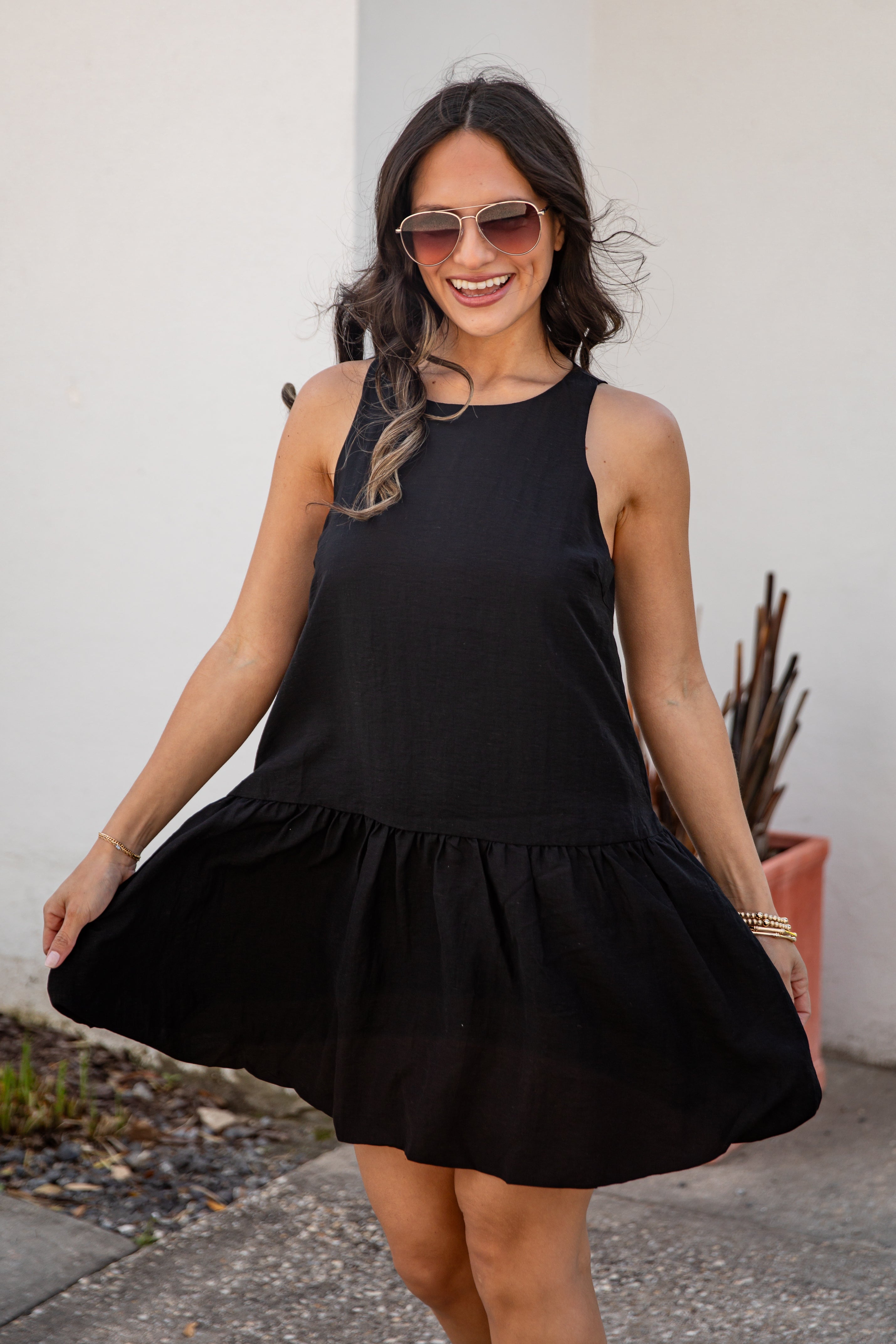 Woman wearing a black dress and sunglasses, standing against a white wall.