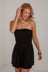 A woman modeling a black strapless mini romper with a ruched bodice and flared skirt, standing against a white wall.