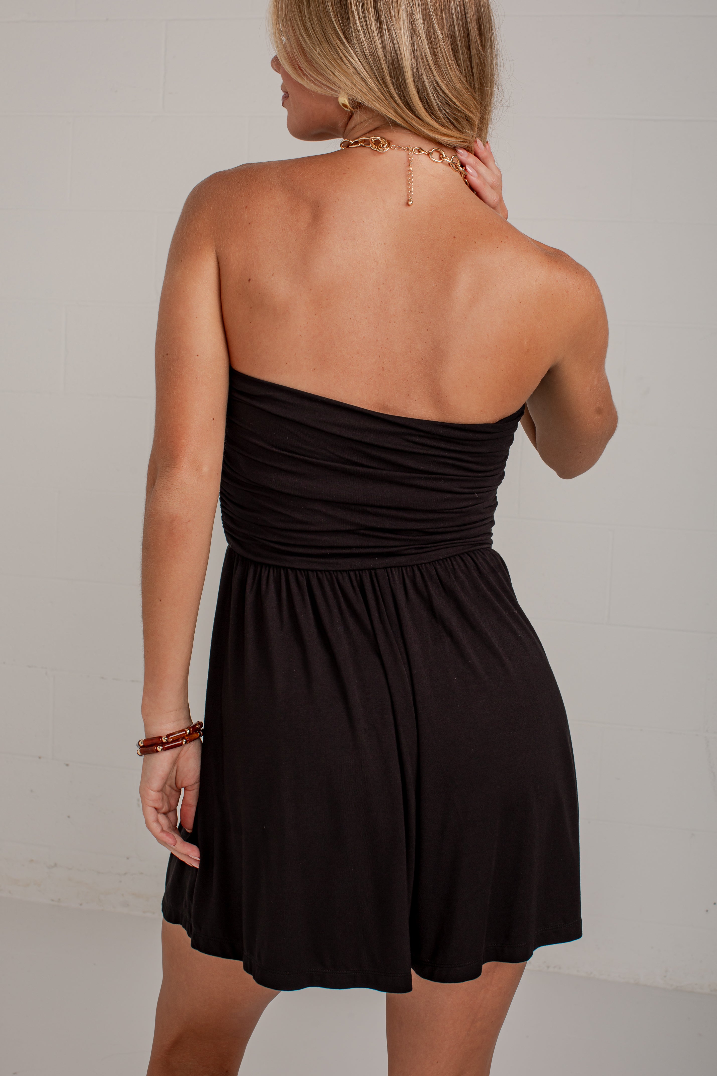 A woman shown from the back wearing a strapless black mini dress with a ruched bodice and flared skirt, photographed against a plain light wall.