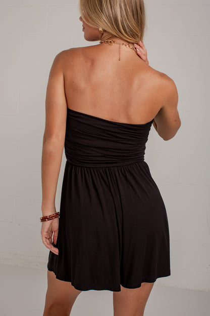 A woman shown from the back wearing a strapless black mini dress with a ruched bodice and flared skirt, photographed against a plain light wall.