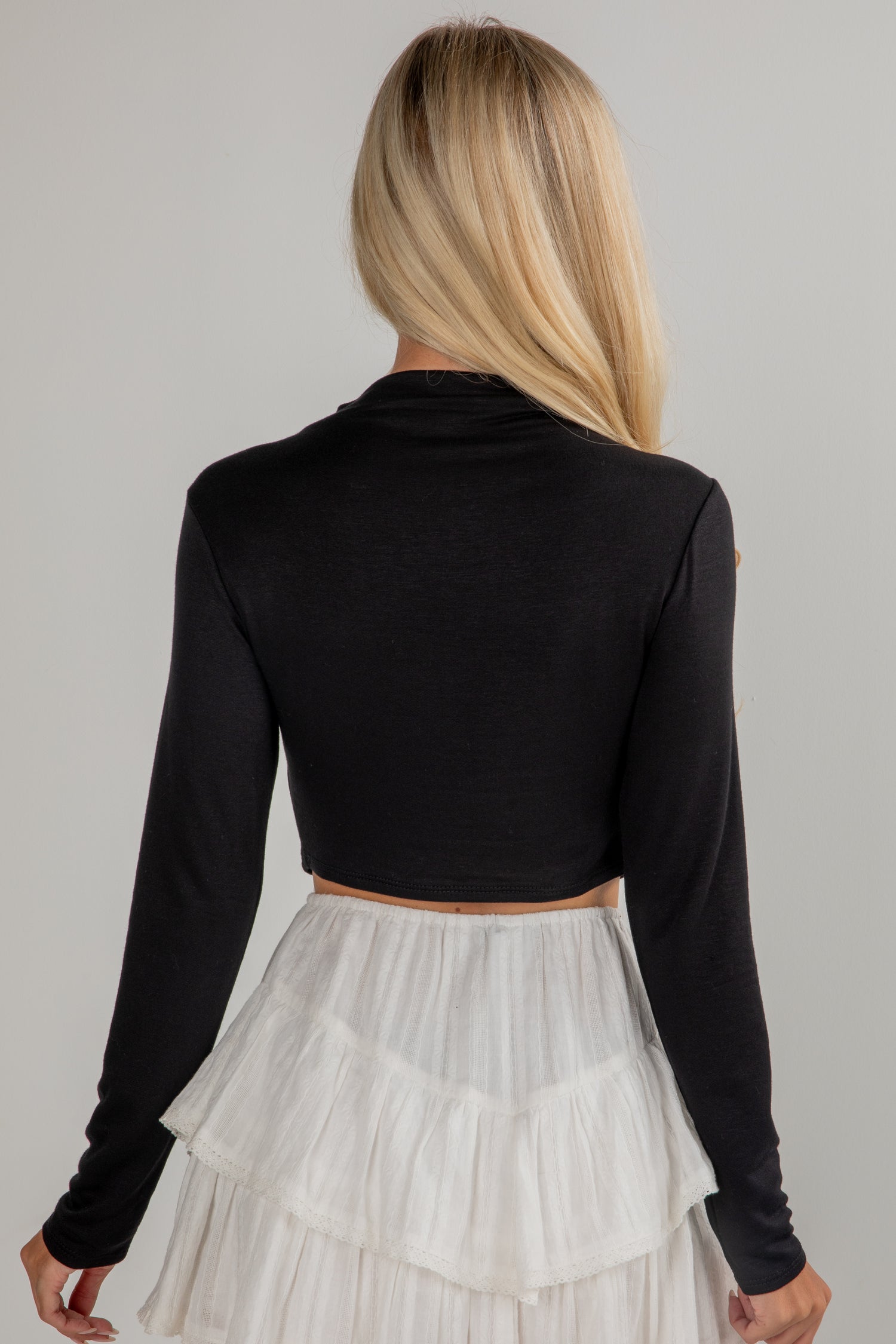 Person wearing a black long-sleeve crop top and white pleated skirt on a gray background