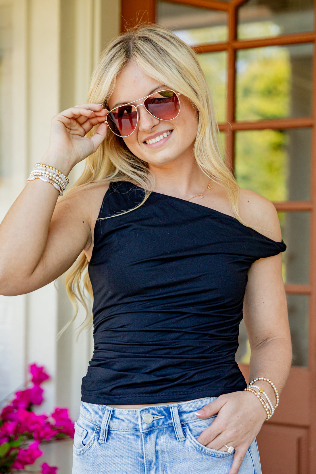 A woman wearing a dark navy blue off-the-shoulder ruched top and light blue jeans, accessorized with gold-framed sunglasses and bracelets, posing in front of a brown wooden door.