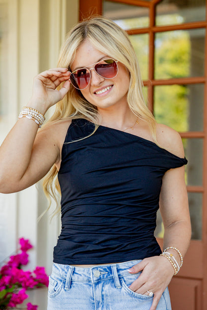 A woman wearing a dark navy blue off-the-shoulder ruched top and light blue jeans, accessorized with gold-framed sunglasses and bracelets, posing in front of a brown wooden door.