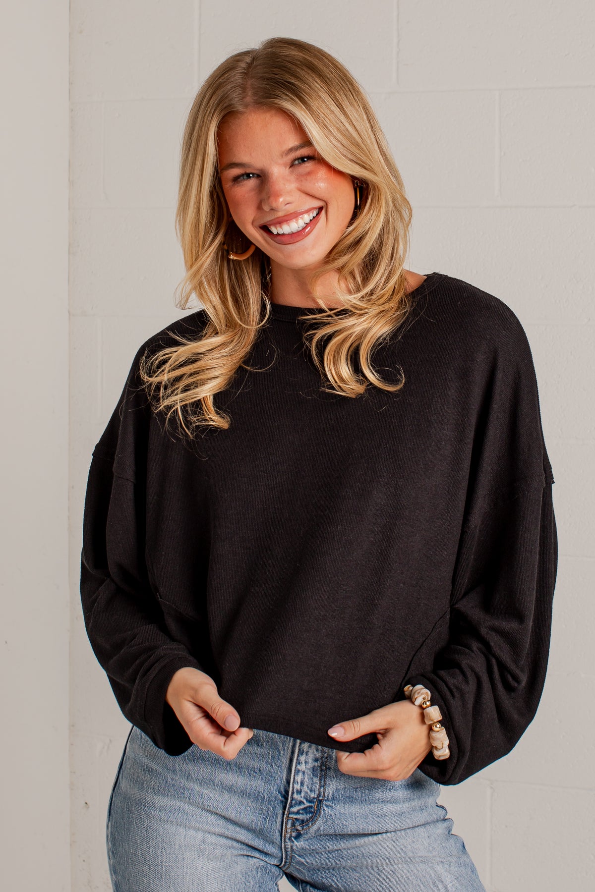 A smiling blonde woman wearing a black long-sleeve crewneck sweatshirt and light blue jeans, standing against a light gray brick wall.
