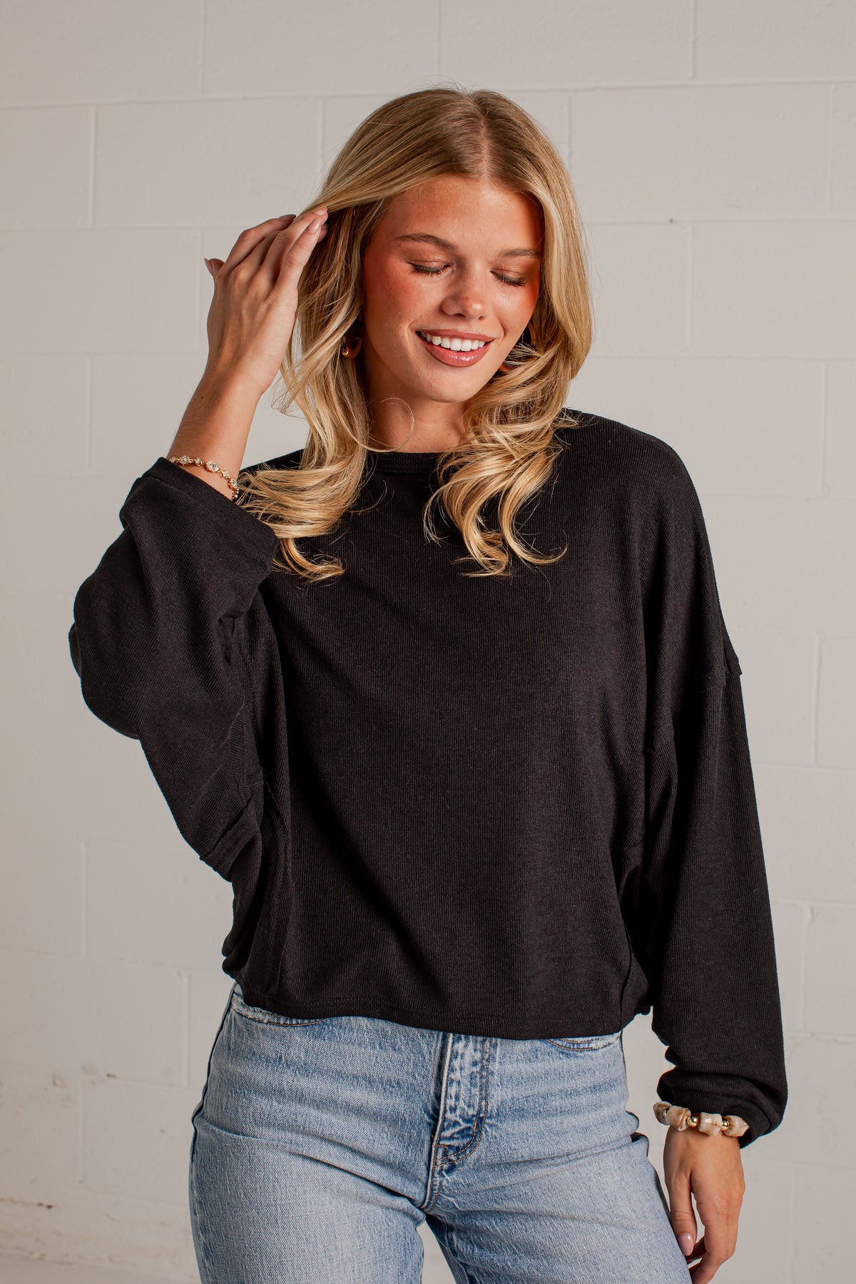 A woman wearing a black long-sleeve crewneck sweatshirt and light wash blue jeans, standing against a white brick wall.